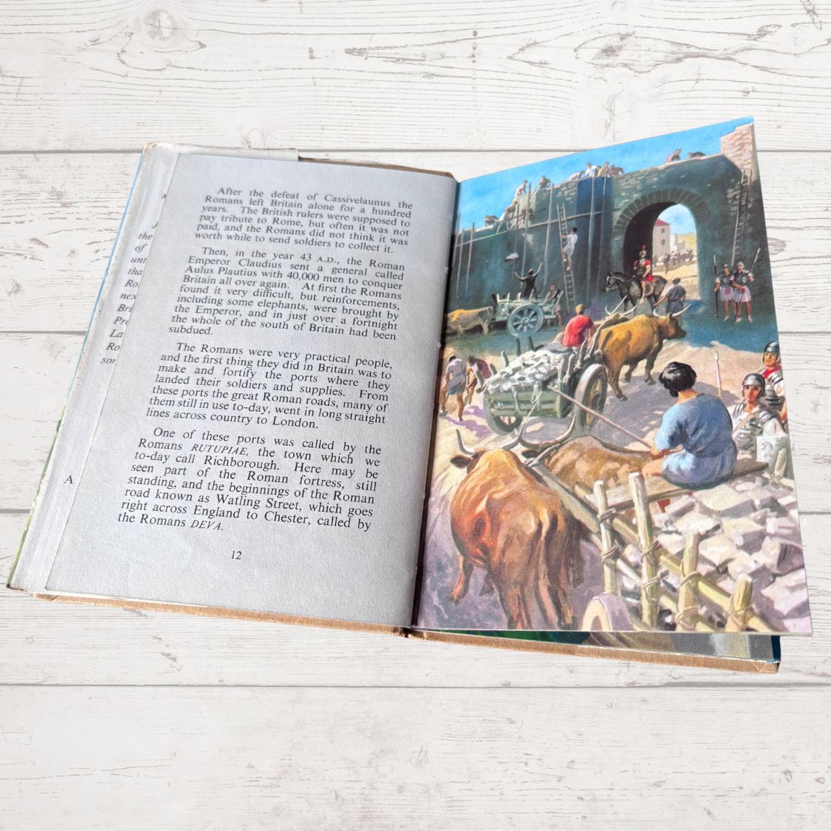 An open vintage book lying flat on a white wooden surface, showing printed text on the left page and a colorful illustration of ancient Roman figures and architecture on the right page.