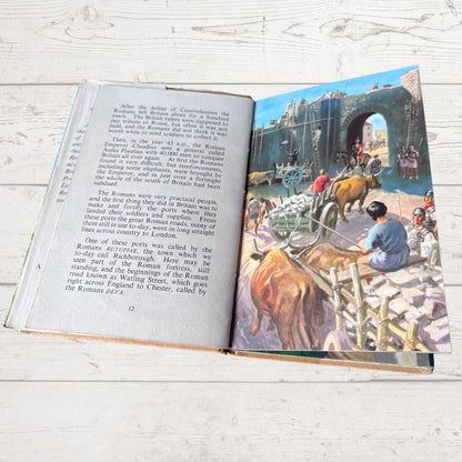 An open vintage book lying flat on a white wooden surface, showing printed text on the left page and a colorful illustration of ancient Roman figures and architecture on the right page.