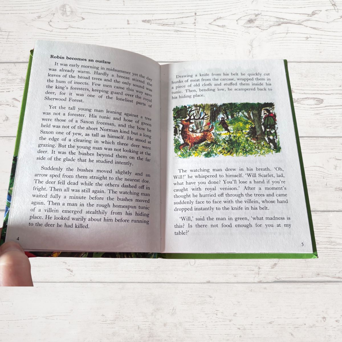 An open book showing two pages of printed text with a green border, photographed on a light wood surface with a hand holding the lower left corner.