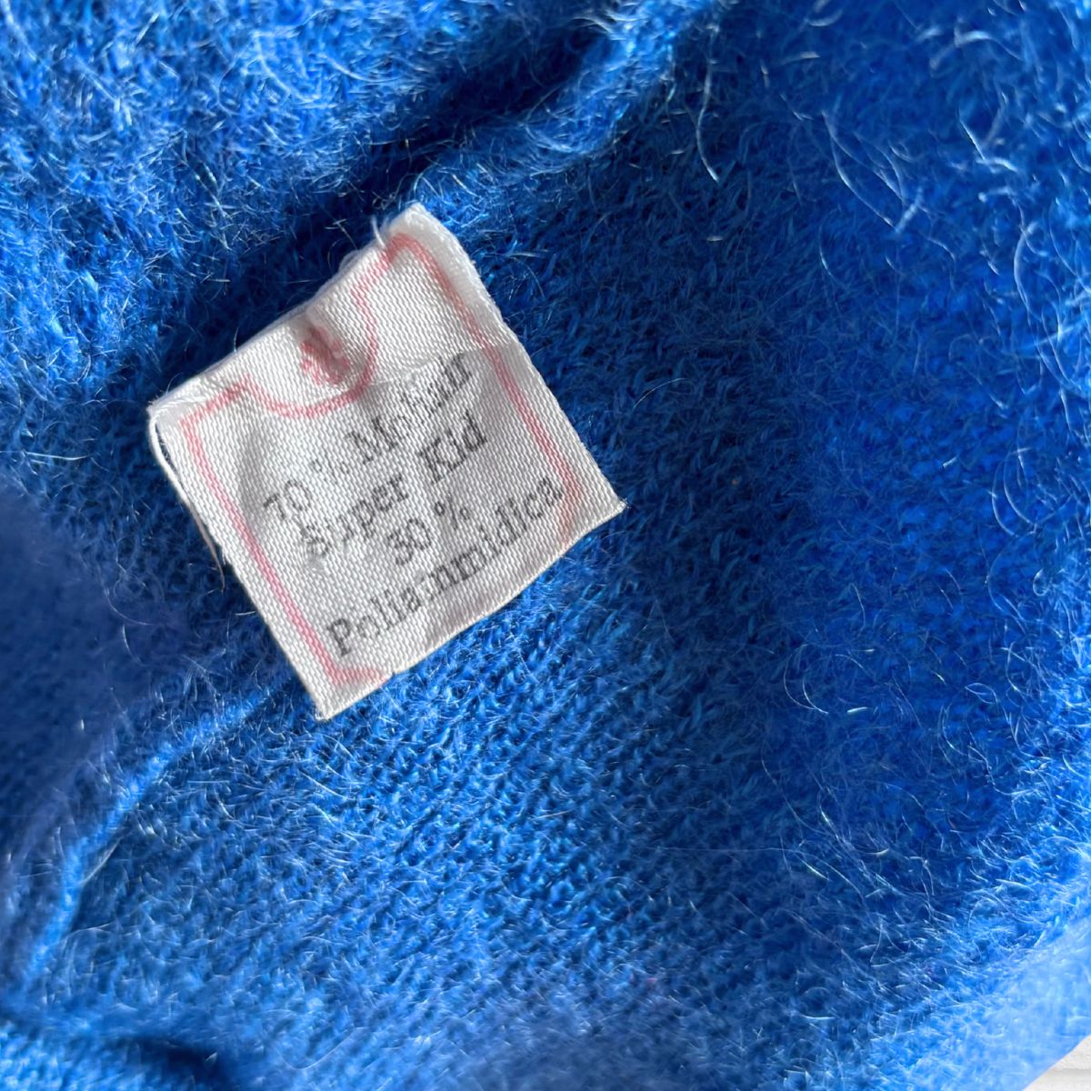A close-up of a bright blue fuzzy knit garment showing a sewn-on fabric label that reads 70% mohair 30% poloAmmidilica 