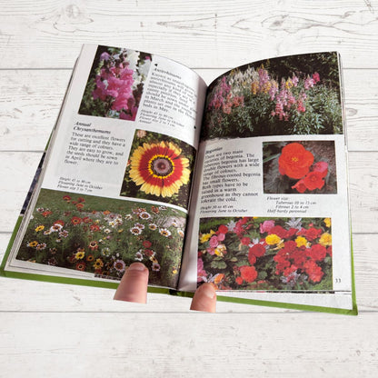 An open gardening book showing colorful flower photographs and text on plant care, held open by two fingers on a white wood surface.