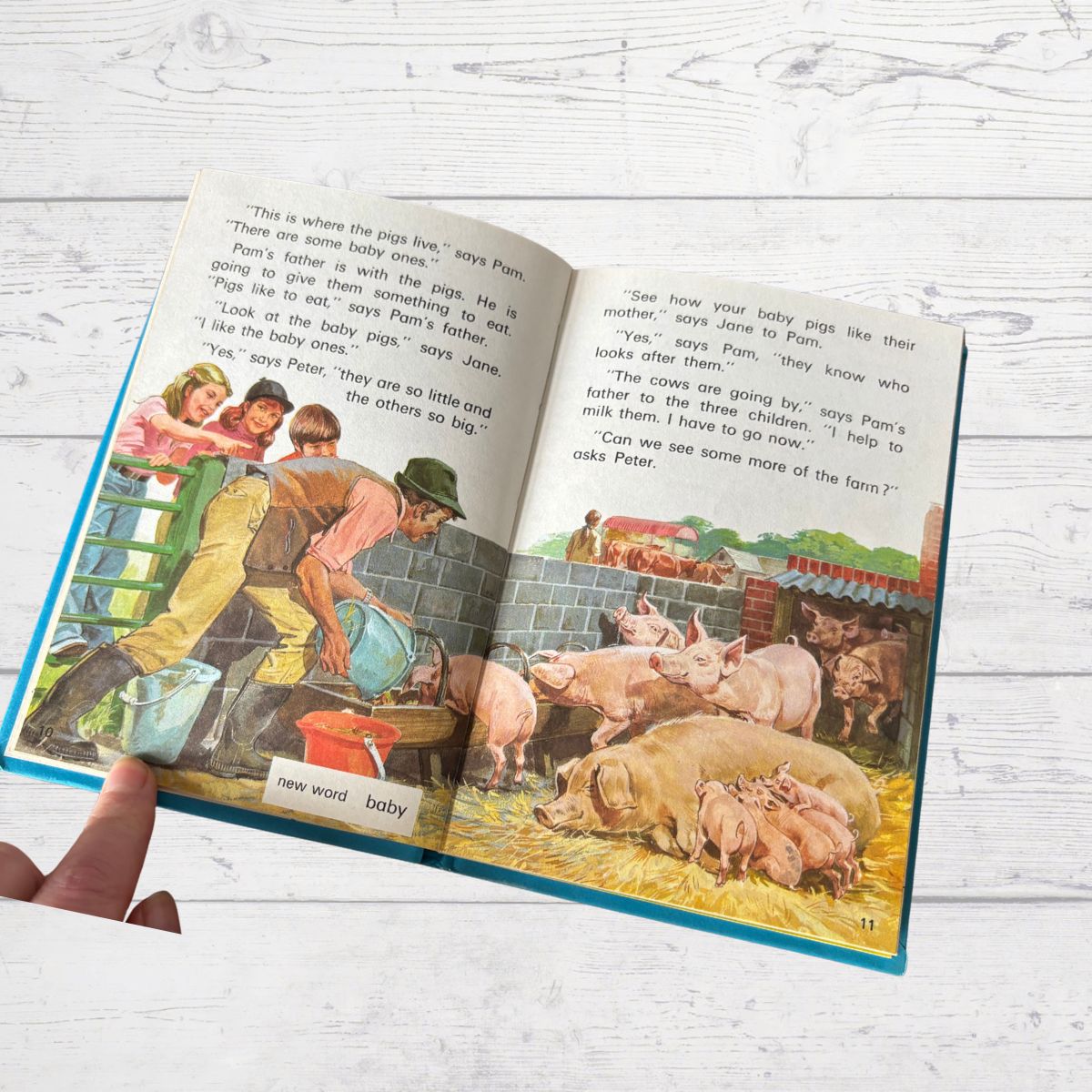 Open book showing a farm scene with pigs and children, held by a hand on a light wooden surface.