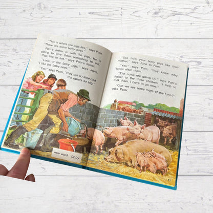 Open book showing a farm scene with pigs and children, held by a hand on a light wooden surface.
