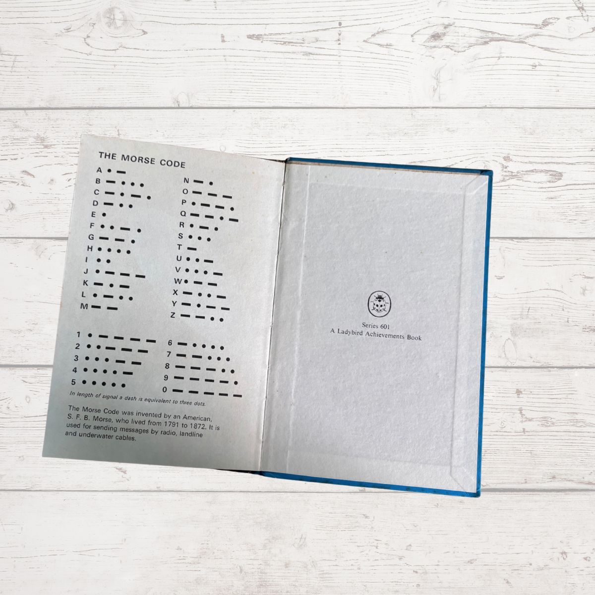 An open vintage book titled 'The Morse Code' with printed black code charts on white pages and a blue cover edge, photographed on a whitewashed wood surface.