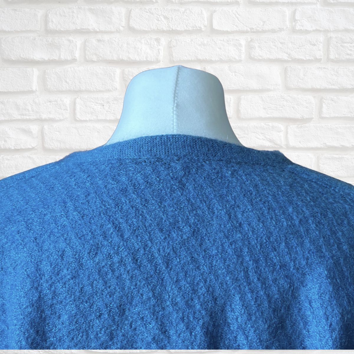 A blue textured knit sweater displayed on a light blue mannequin torso, shown from the back against a white brick wall background.