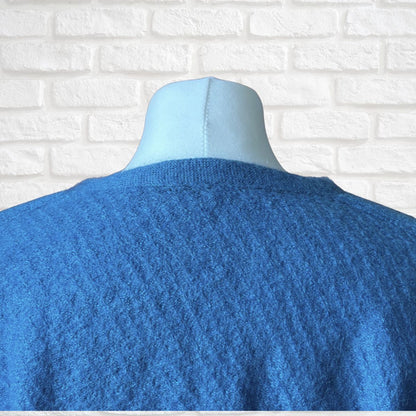 A blue textured knit sweater displayed on a light blue mannequin torso, shown from the back against a white brick wall background.
