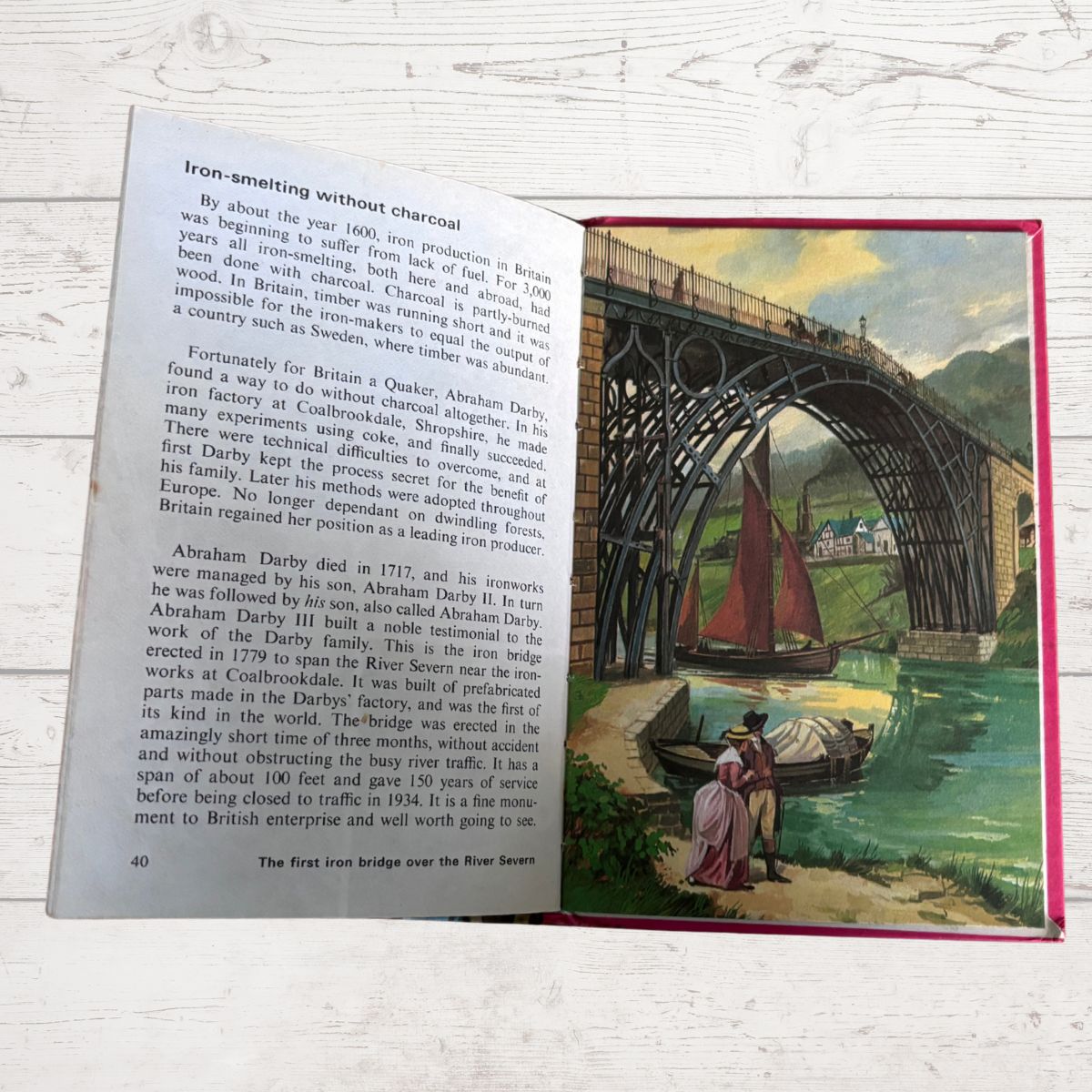 An open vintage book or notebook lying on a white wood surface, showing printed text on one page and a colorful illustration on the other. The illustration depicts a landscape with a bridge, water, and a figure in a red coat.