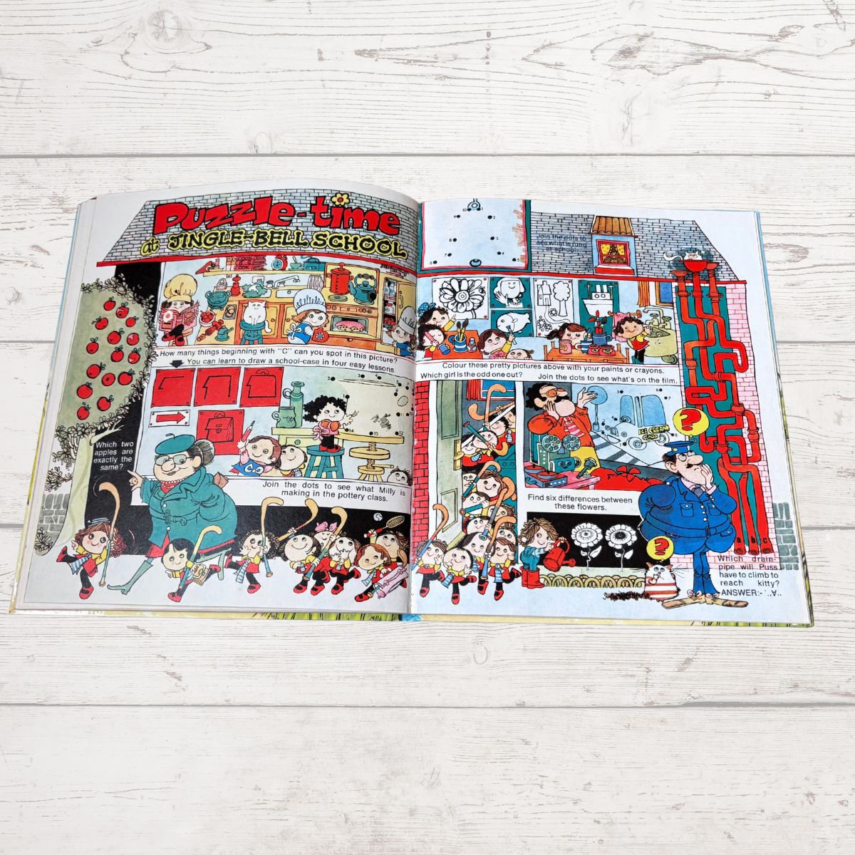 An open vintage children's puzzle book spread on a white wood plank surface, featuring colorful comic-style illustrations and the title 'Puzzle-time: A Single-Ball School.'.