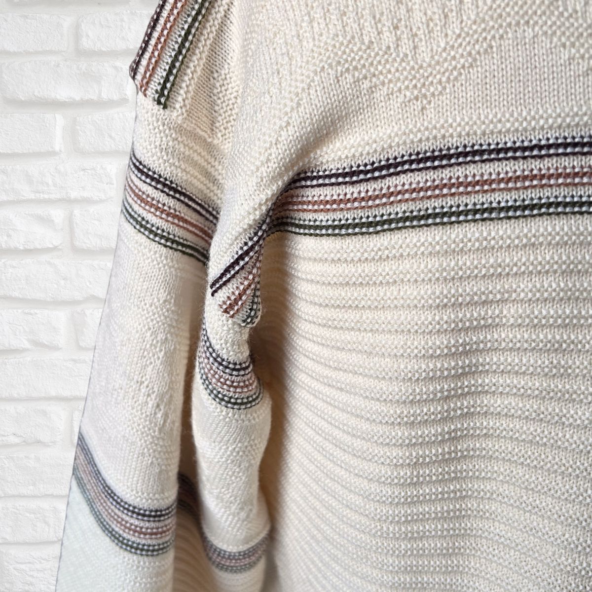 Close-up of a cream-colored knit sweater with horizontal stripes ,shown against a white brick wall background 