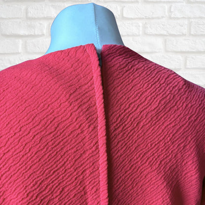 A close-up of the back of a textured hot pink dress displayed on a light blue mannequin torso, set against a white brick wall background.