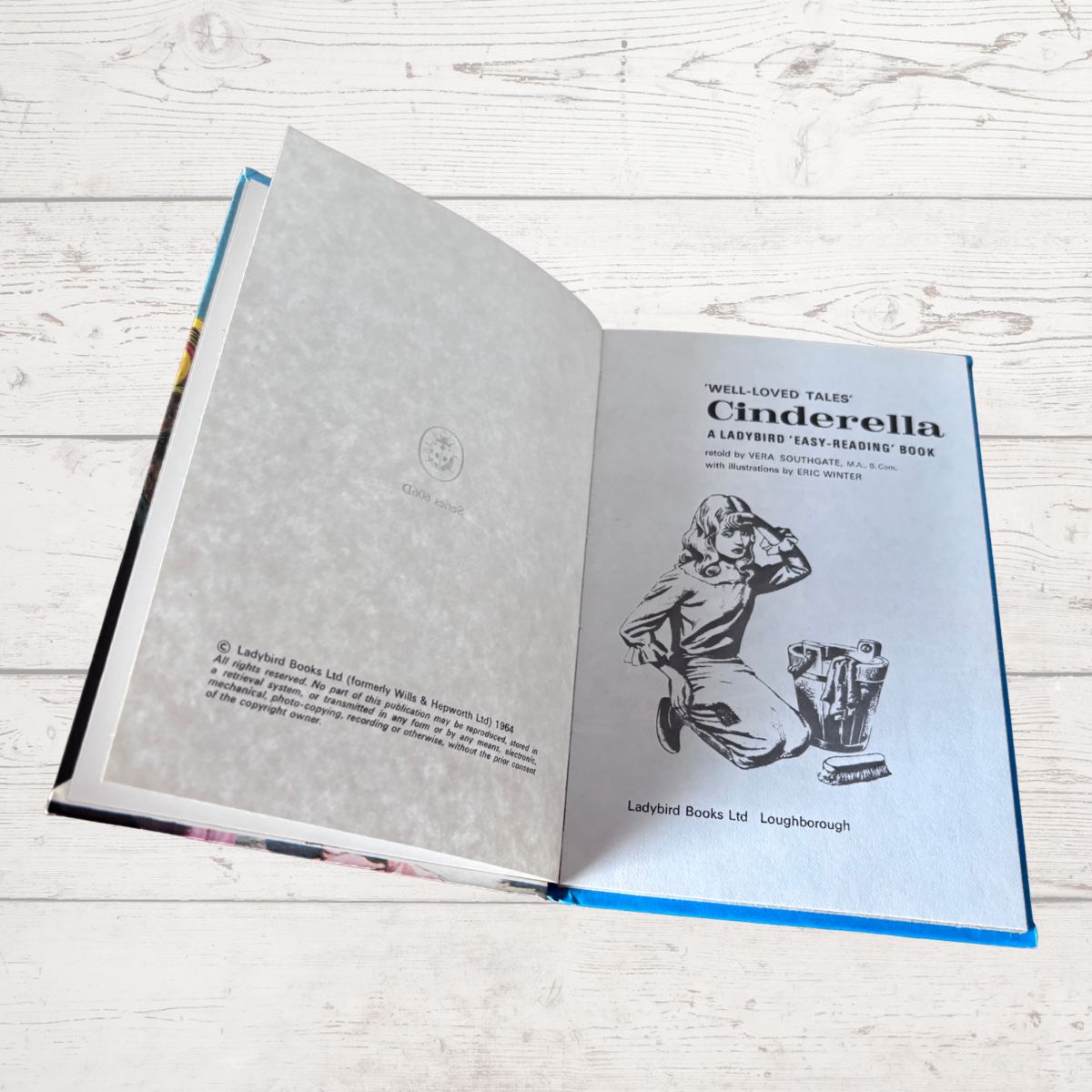 An open hardcover book titled 'Cinderella' lying on a light whitewashed wooden surface. The left page is blank with faint decorative patterning, and the right page shows the title, author text, and a black-and-white illustration of Cinderella kneeling by a mirror.