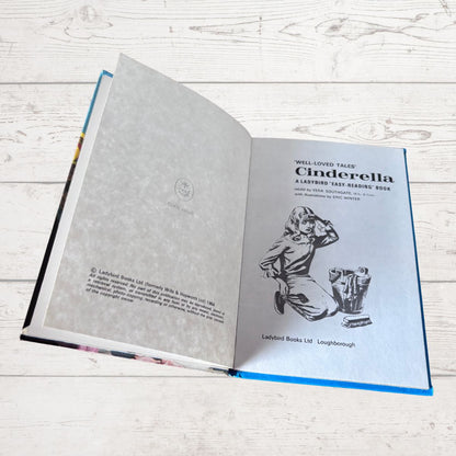 An open hardcover book titled 'Cinderella' lying on a light whitewashed wooden surface. The left page is blank with faint decorative patterning, and the right page shows the title, author text, and a black-and-white illustration of Cinderella kneeling by a mirror.