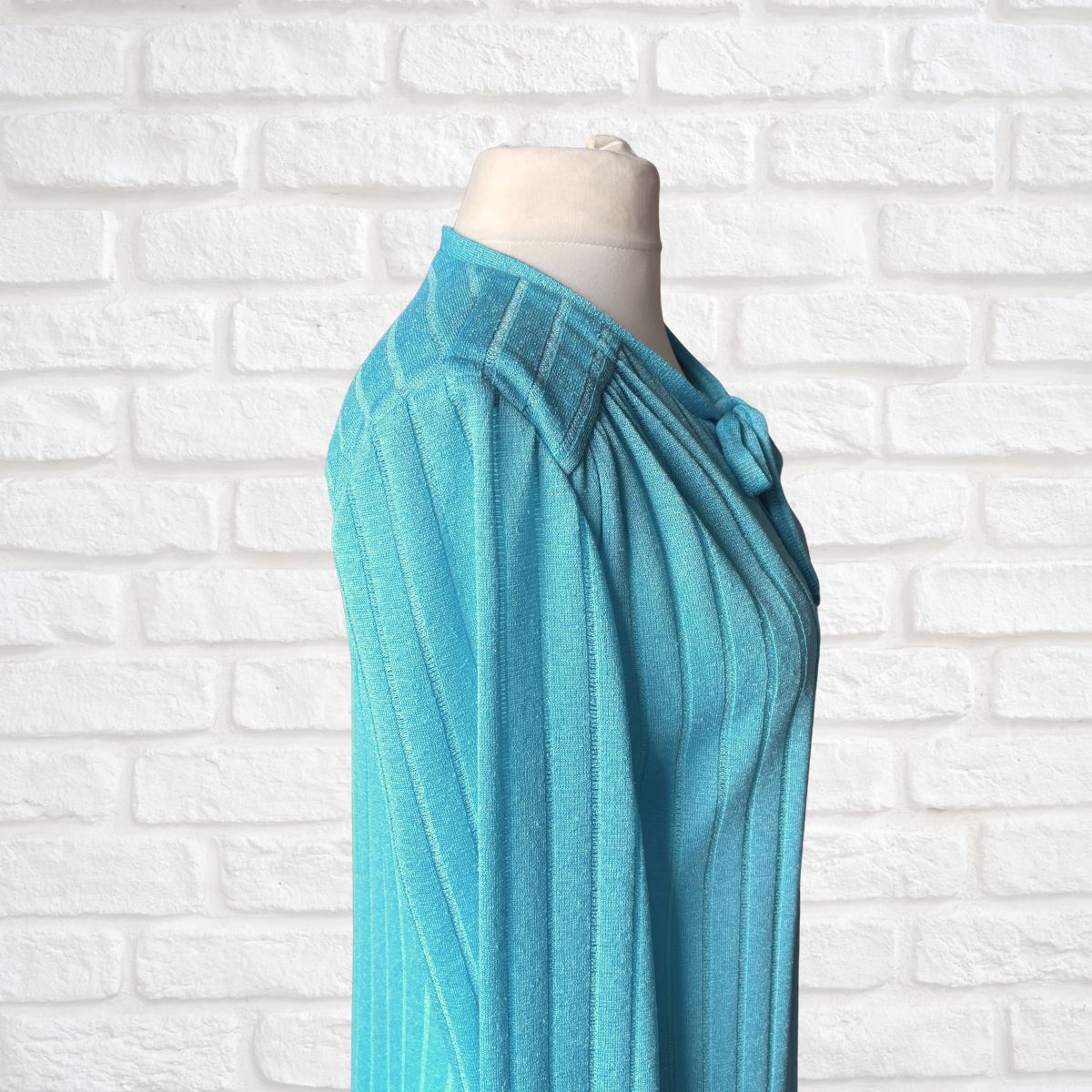 A turquoise blue knit displayed on a cream mannequin torso against a white painted brick wall background.