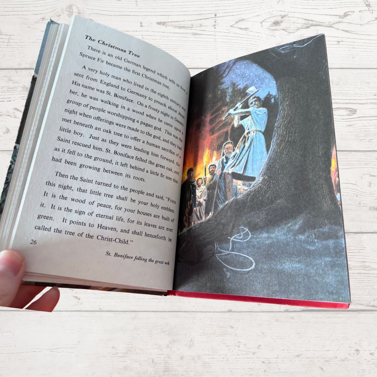 An open children's book titled 'The Christmas Tree' being held by a hand, showing printed text on one page and a colorful illustration of a tree with a glowing Christmas tree on the other, set against a light wood background.