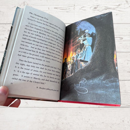 An open children's book titled 'The Christmas Tree' being held by a hand, showing printed text on one page and a colorful illustration of a tree with a glowing Christmas tree on the other, set against a light wood background.
