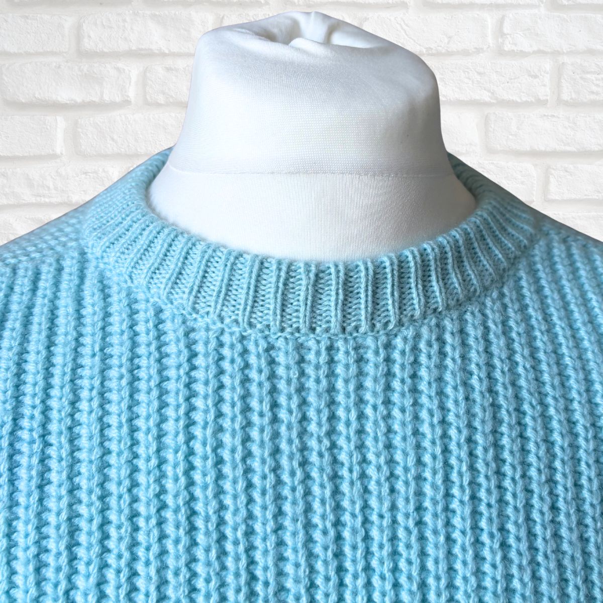 A light blue ribbed knit sweater displayed on a white mannequin torso against a white brick wall background.