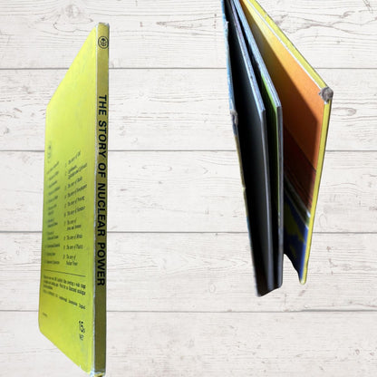 A vintage hardcover book titled 'The Story of Nuclear Power' shown standing upright on its spine, with its pages fanned out to reveal multiple colored interior covers, set against a light wood plank background.
