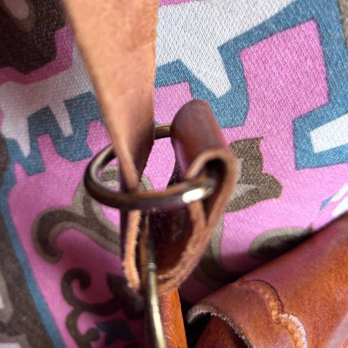 Close-up detail of a brown leather strap attached to a metal ring and loop, resting against a pink, blue, and gray patterned fabric background.