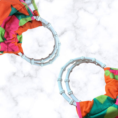 A close-up of a  fabric bag with a bright multicolour pattern in pink, orange, green, and teal, featuring a white plastic  bamboo-style handle on a white marble background.