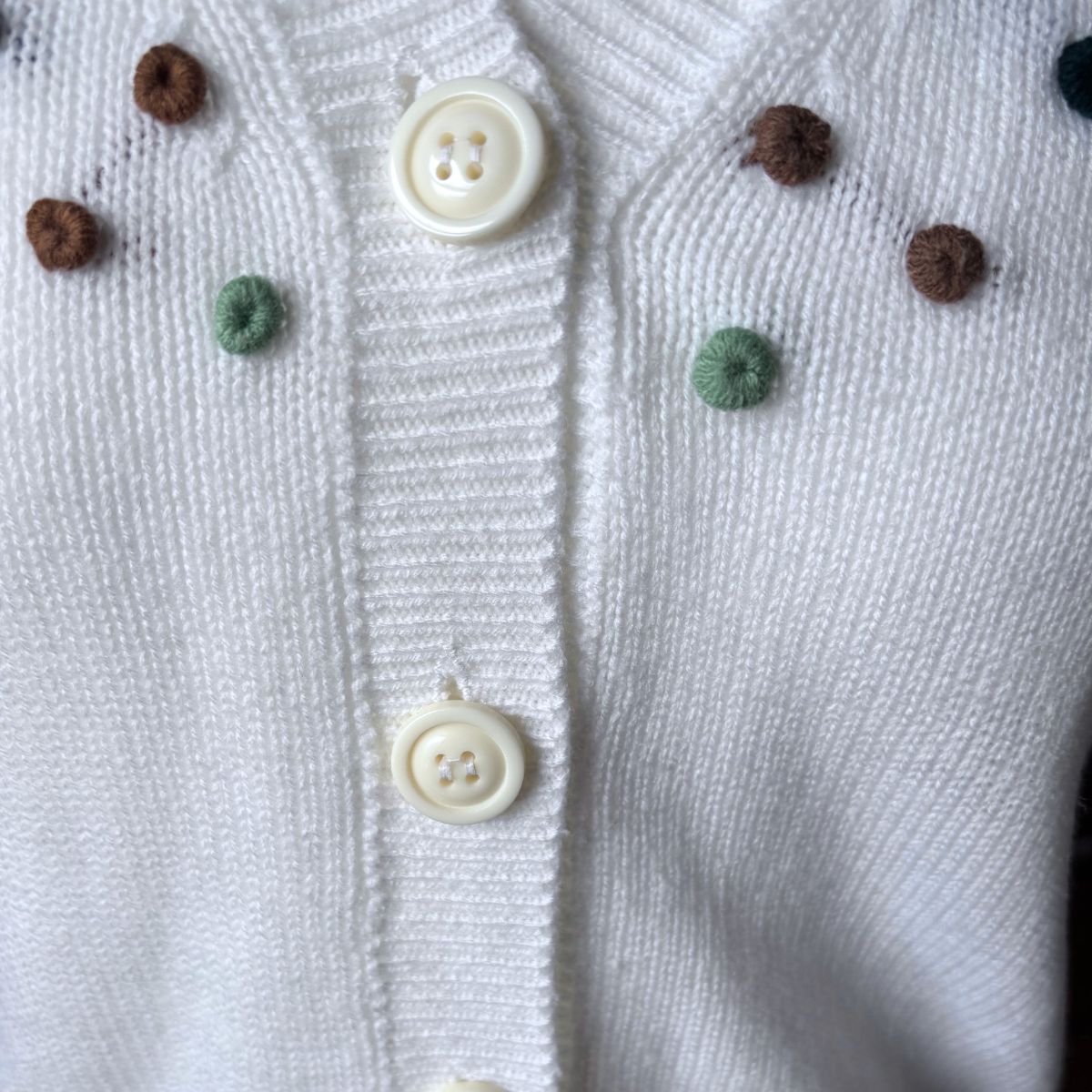 Close-up of a white knit cardigan with cream buttons and small decorative pom-pom accents in brown and mint green.