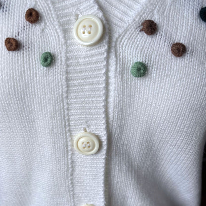 Close-up of a white knit cardigan with cream buttons and small decorative pom-pom accents in brown and mint green.