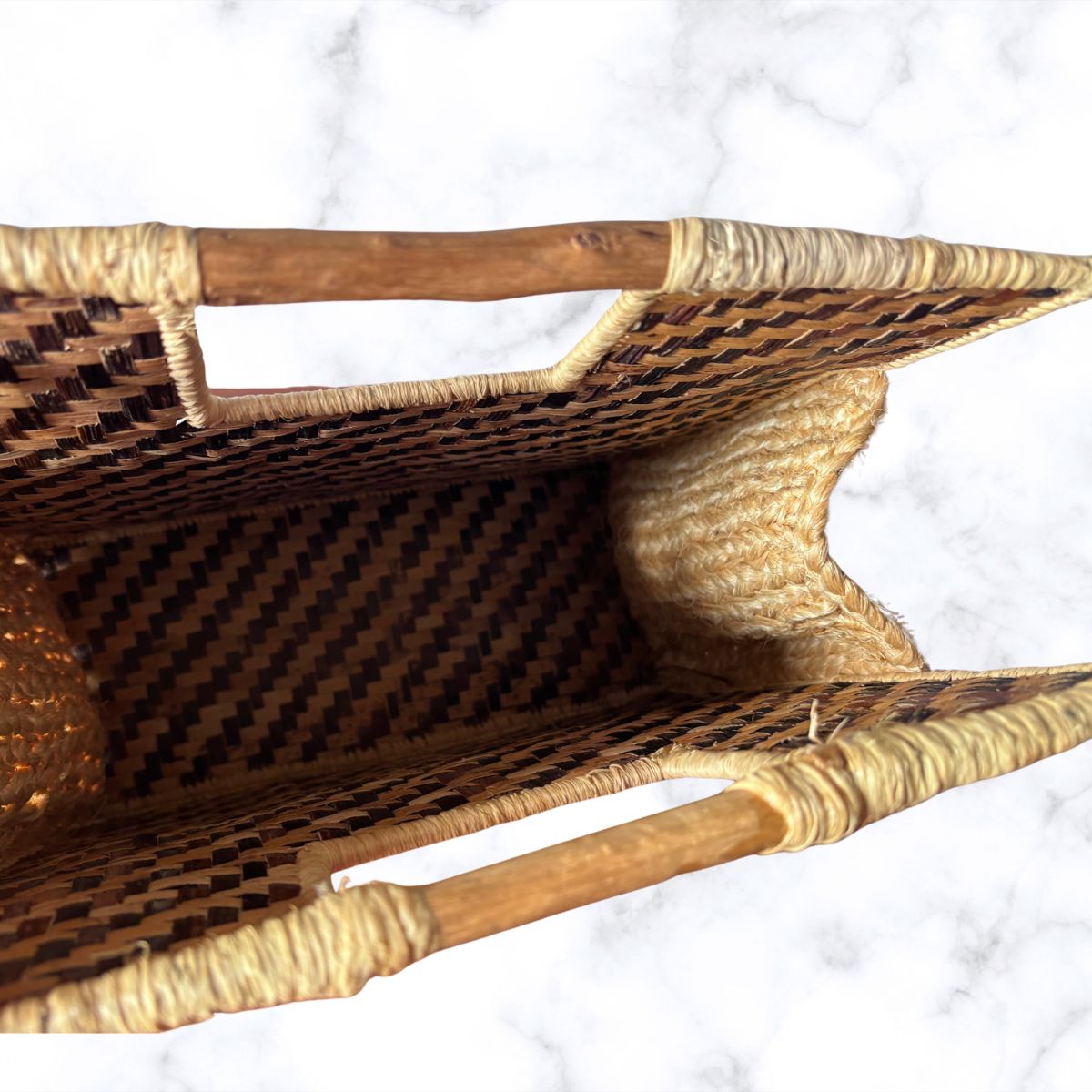 A close-up top-down view of a woven rattan or seagrass basket with a diamond-patterned weave, natural fibre trim, and wooden handles, set against a white marble background.