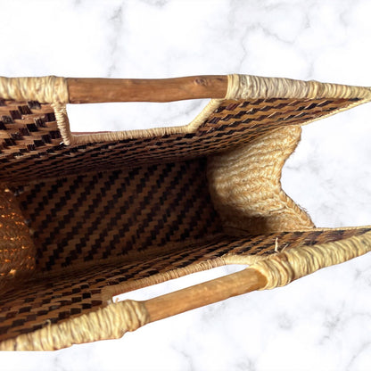 A close-up top-down view of a woven rattan or seagrass basket with a diamond-patterned weave, natural fibre trim, and wooden handles, set against a white marble background.