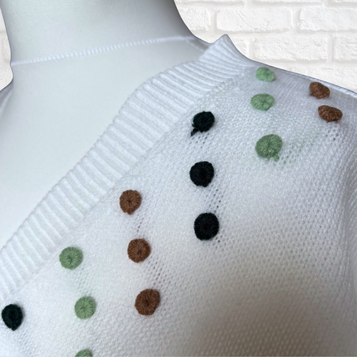 White knit sweater with small multicolor pom-pom dots in black, brown, and green, displayed on a white mannequin torso against a white brick wall background.