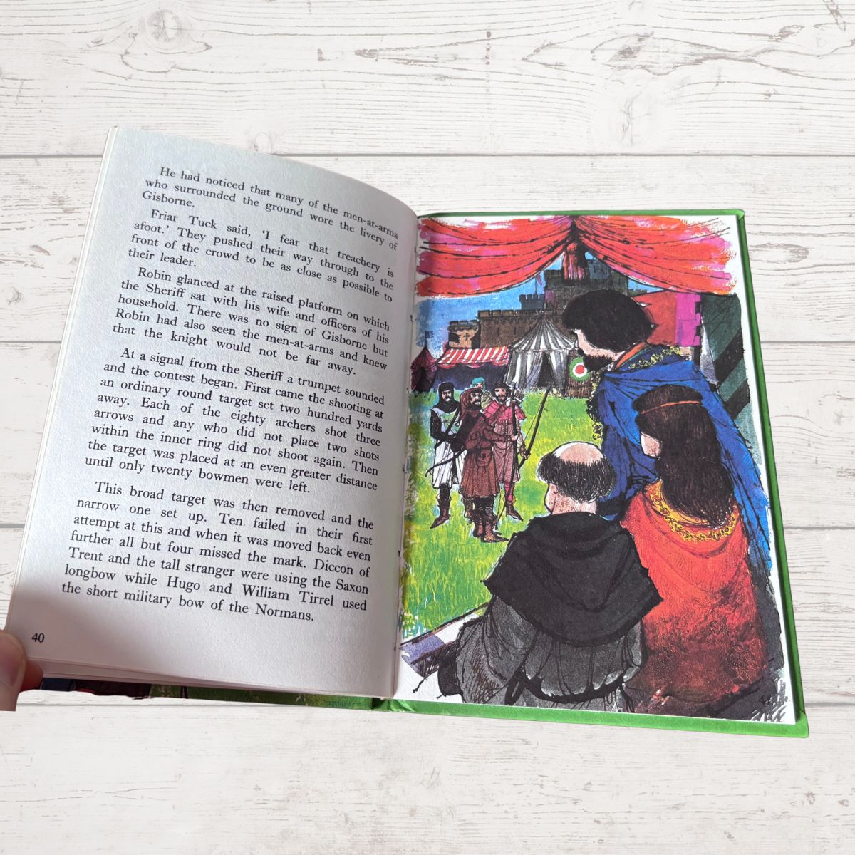 An open children's book lying on a white wood surface, showing printed text on the left page and a colorful illustration on the right page. A hand is visible holding the left edge of the book.