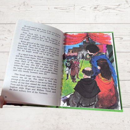 An open children's book lying on a white wood surface, showing printed text on the left page and a colorful illustration on the right page. A hand is visible holding the left edge of the book.