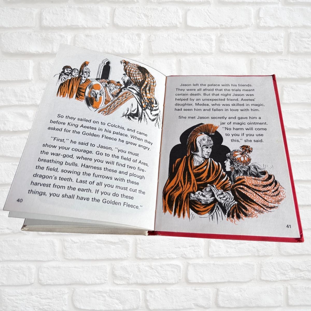 An open children's book lying on a white brick surface, showing two illustrated pages with fantasy artwork and printed text. The left page has a black-and-white illustration with a small colored accent, and the right page has a larger black-and-white portrait with orange and brown flowing hair details.