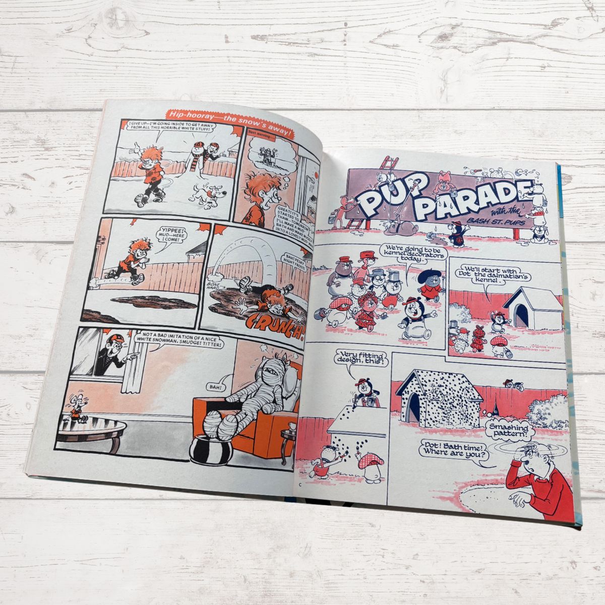 An open comic book or graphic novel lying on a white wooden surface. The pages show a cartoon strip titled 'Pup Parade' with multiple panels, speech bubbles, and illustrated characters in red, black, white, and orange tones.