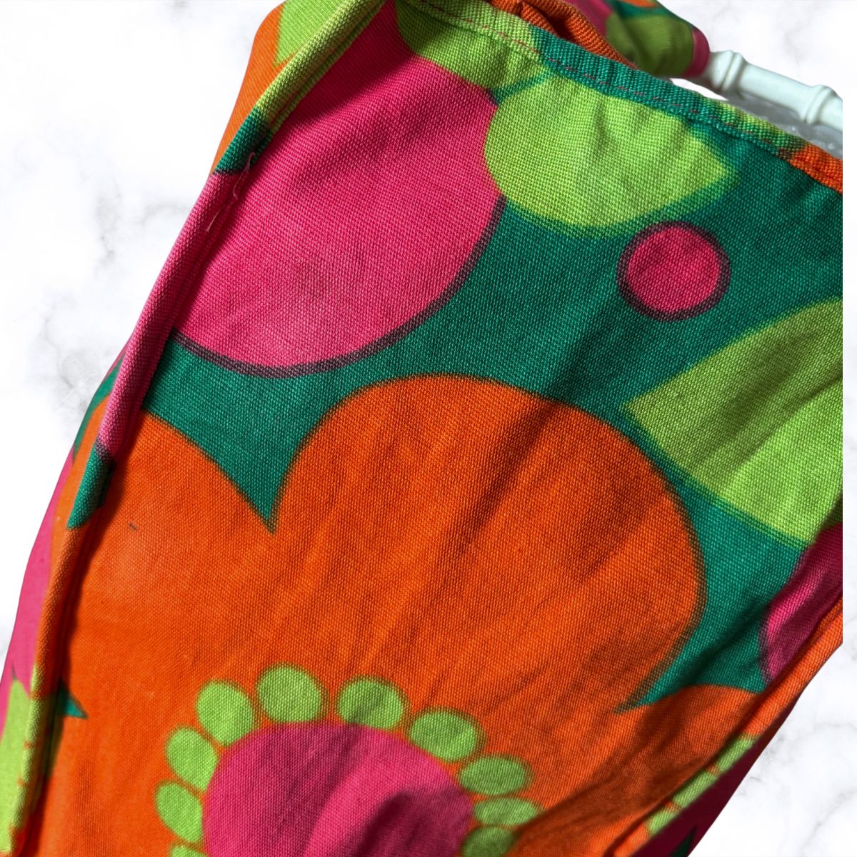 A close-up of a colourful patterned bag  featuring a retro floral design in orange, green, pink, and yellow on a white marble background.