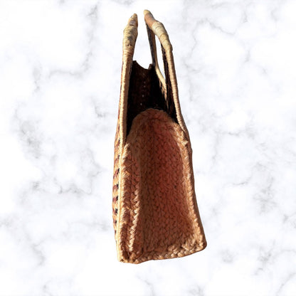 A woven straw tote bag with two handles, shown from a top-down view on a white marble background.