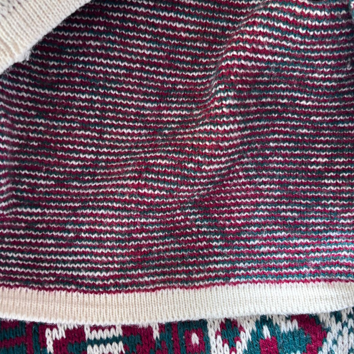 Close-up of a knitted fabric with horizontal stripes in burgundy, cream, and dark gray, with a ribbed cream border and a patterned section at the bottom.