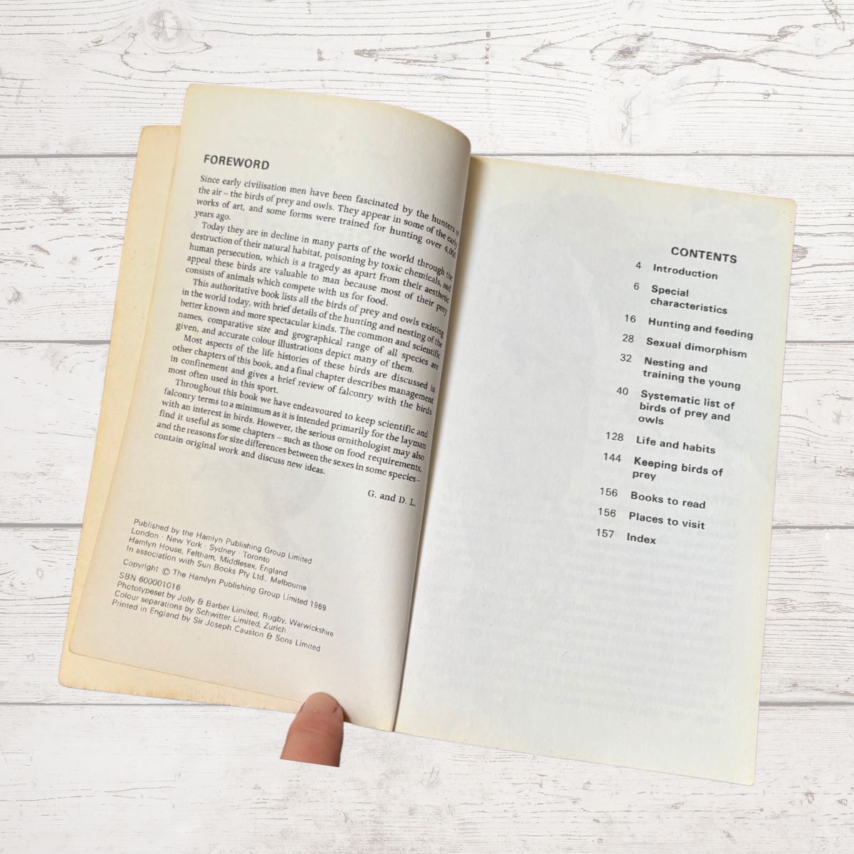 An open vintage book showing the foreword and contents pages, held by a hand against a whitewashed wooden background.