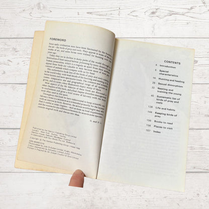 An open vintage book showing the foreword and contents pages, held by a hand against a whitewashed wooden background.