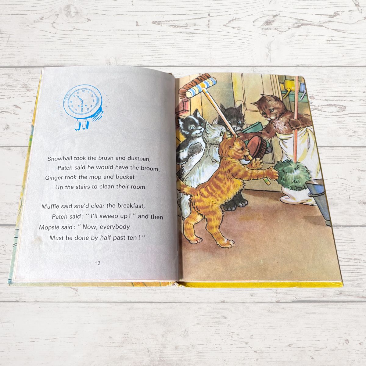 An open children's book lying on a white wood surface, showing a printed story with text on the left page and a colorful illustration of cats cleaning a room on the right page.