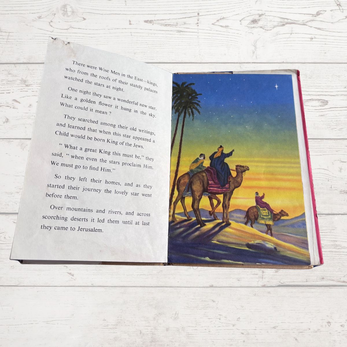 Open book with a colorful illustration of three people on camels against a sunset sky, on a wooden surface.
