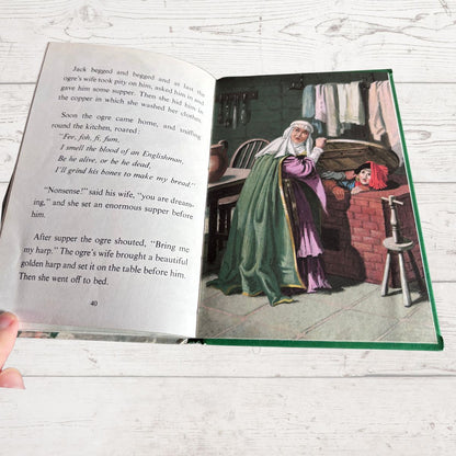 An open children's book lying on a white wood surface, showing printed text on the left page and a colorful illustration on the right page of an elderly woman in a green dress standing by a window.