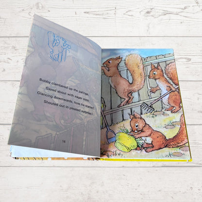 An open children's picture book lying on a light wood surface, showing illustrated squirrels by a fence on one page and text on the other.