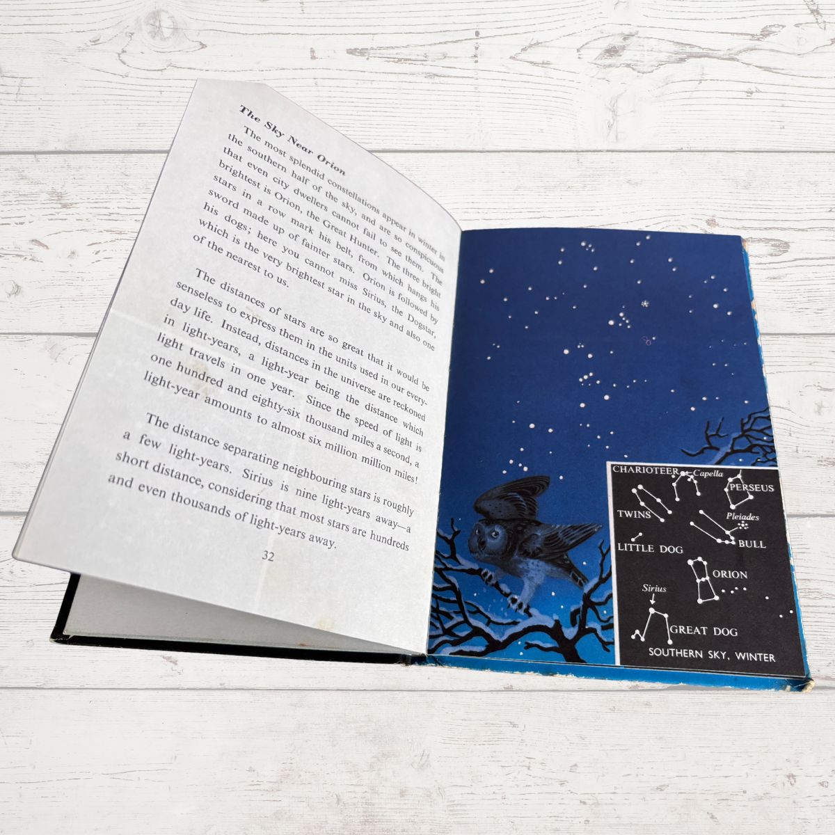 An open book showing a page about the night sky and a colorful illustration of a starry night with a constellation diagram, placed on a white wood surface.