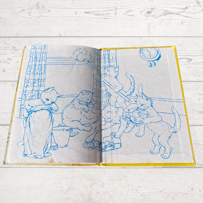 An open vintage children's book lying flat on a white wooden surface. The pages feature blue line drawings of animals, including a cat, dog, and rabbit, performing chores, with a yellow border visible around the pages.