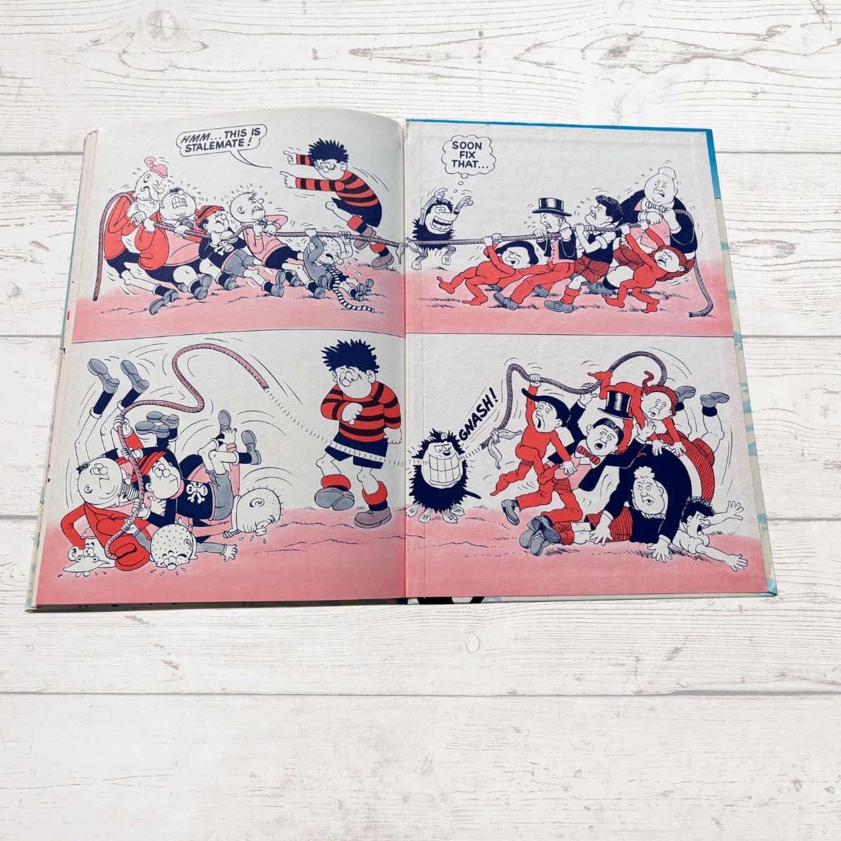 An open vintage comic book laid flat on a white wood surface, showing four-panel cartoon scenes with red, black, white, and pink tones.
