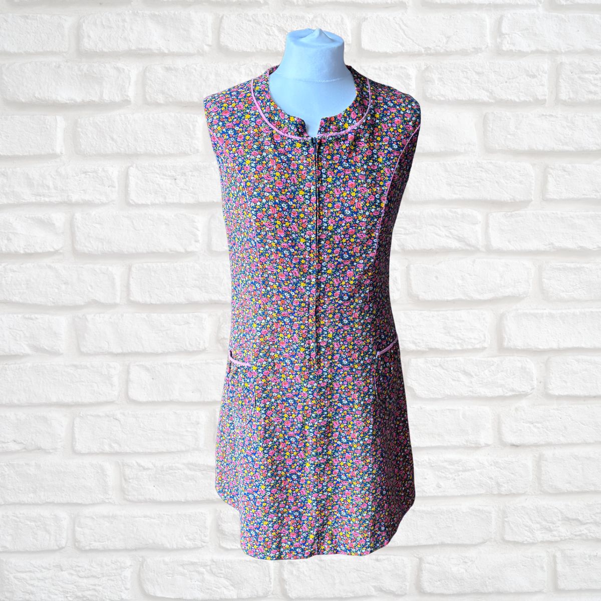 A sleeveless zip-front dress with a dense multicolour  floral print, displayed on a light blue mannequin torso against a white brick wall background.