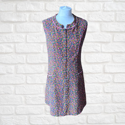 A sleeveless zip-front dress with a dense multicolour  floral print, displayed on a light blue mannequin torso against a white brick wall background.