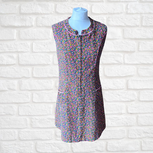 A sleeveless zip-front dress with a dense multicolour  floral print, displayed on a light blue mannequin torso against a white brick wall background.