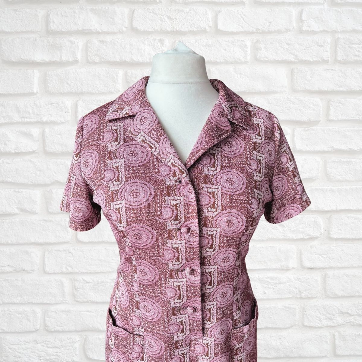 A short-sleeve collared button-front dress with an allover pink, mauve, and off-white geometric pattern, displayed on a white mannequin torso against a white brick wall background.