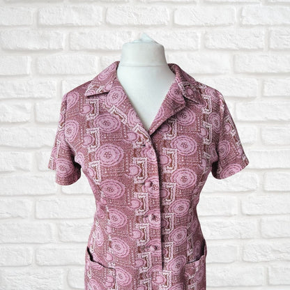 A short-sleeve collared button-front dress with an allover pink, mauve, and off-white geometric pattern, displayed on a white mannequin torso against a white brick wall background.