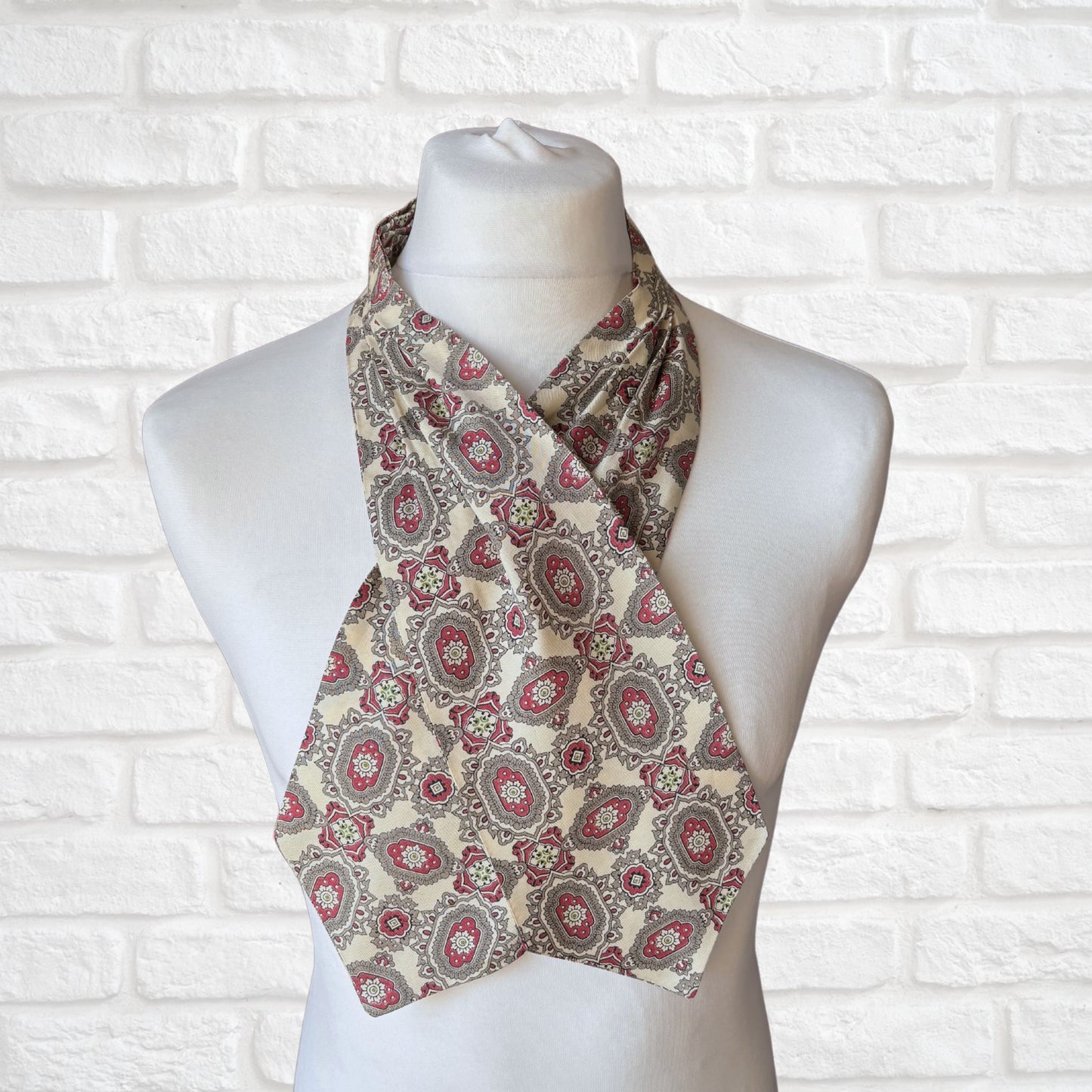 A vintage 60s mod style cream cravat with a paisley print featuring baroque style red, grey, and green patterns, displayed on a mannequin against a white brick wall background.